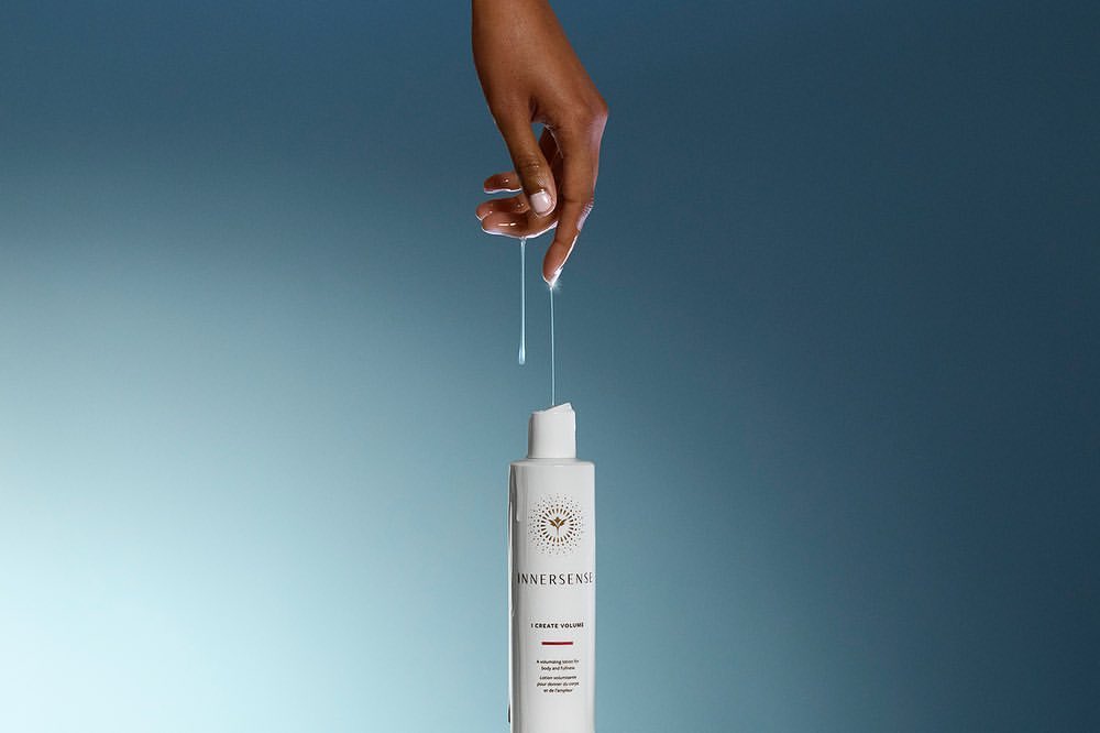 An adult hand touches clear liquid dripping onto a white Innnersense hair product bottle against a blended light blue background.