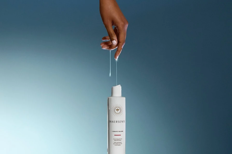 An adult hand touches clear liquid dripping onto a white Innnersense hair product bottle against a blended light blue background.