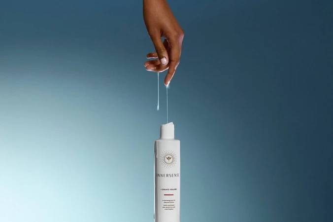 An adult hand touches clear liquid dripping onto a white Innnersense hair product bottle against a blended light blue background.