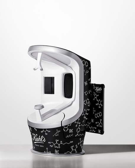Kiehl's skin scanner with a white and silver finish, adorned with molecule patterns, stands on a white surface against a white backdrop.
