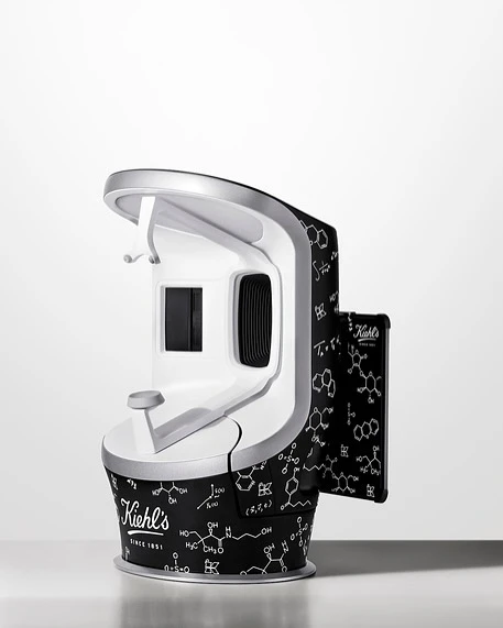 Kiehl's skin scanner with a white and silver finish, adorned with molecule patterns, stands on a white surface against a white backdrop.