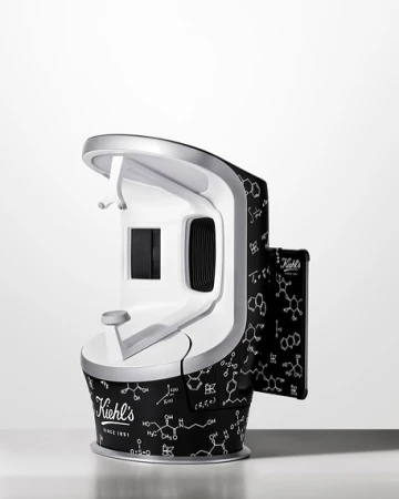 Kiehl's skin scanner with a white and silver finish, adorned with molecule patterns, stands on a white surface against a white backdrop.