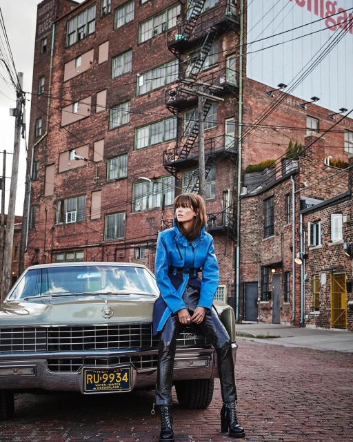 A stylish woman in a blue jacket sits on the hood of a classic car in an urban setting, featuring a brick building backdrop.