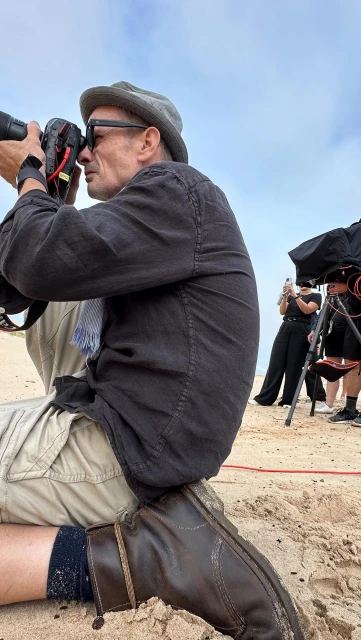 Candid view of a photographer in hat and dark jacket kneeling on a sandy beach with camera gear, capturing outdoor scenes with natural light.