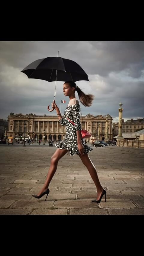 Fashionable woman in a floral dress confidently strides through a city square under an umbrella, capturing a dynamic street style.