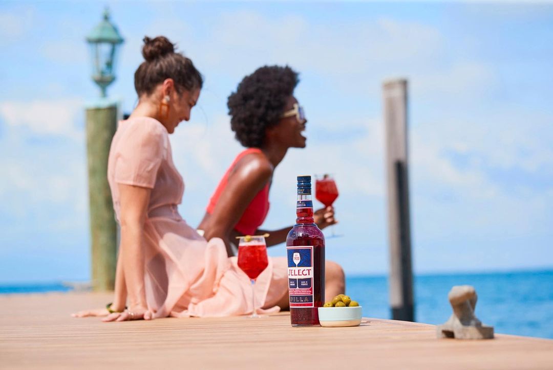 Two women enjoy cocktails on a sunny dock next to a bottle of Select Aperitivo, with the ocean and sky visible in the background.