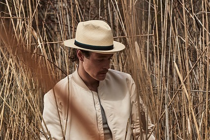 A fair-skinned adult male in a cream-colored bomber jacket and fedora, set against a background of dry reeds on an overcast day.