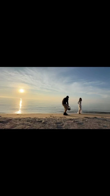 Ocean sunset photoshoot with a woman in a white dress and a photographer on a sandy beach against a backdrop of water and sky.