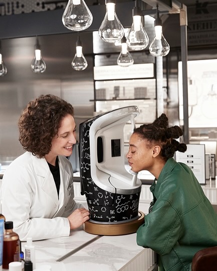 Woman using a Kiehl's skin analysis machine as a skincare specialist looks on, indoors with hanging pendant lights.