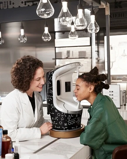 Woman using a Kiehl's skin analysis machine as a skincare specialist looks on, indoors with hanging pendant lights.