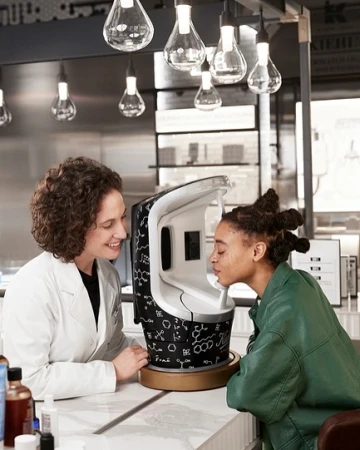 Woman using a Kiehl's skin analysis machine as a skincare specialist looks on, indoors with hanging pendant lights.