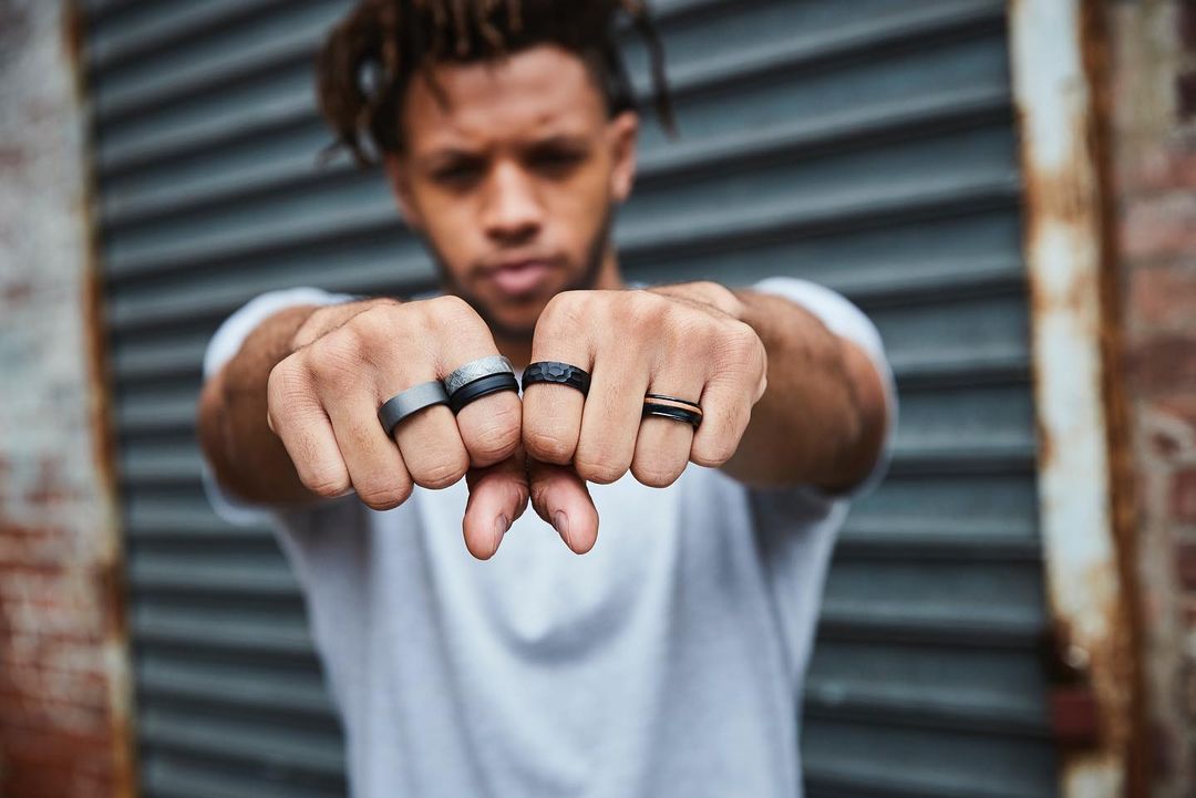 Close-up of a man displaying an assortment of rings on his fingers, with a blurred background of a gray metal wall.