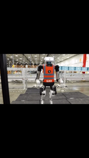Robot stands in warehouse aisle wearing orange reflective vest. Shelves and automated systems fill the background.