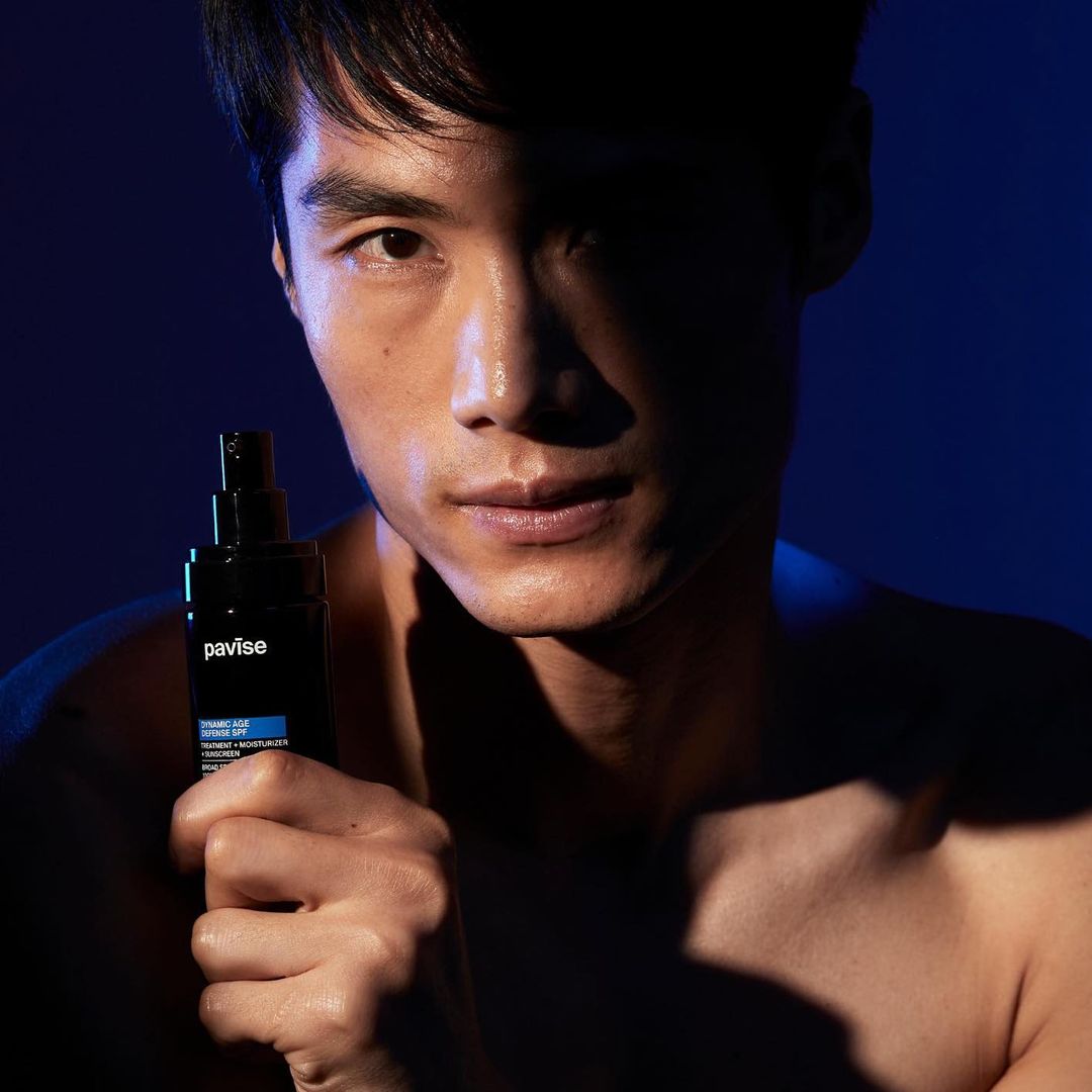 Asian male poses indoors with Pavise Dynamic Milk Defense SPF. He is holding the black and blue bottle with the brand name visible.