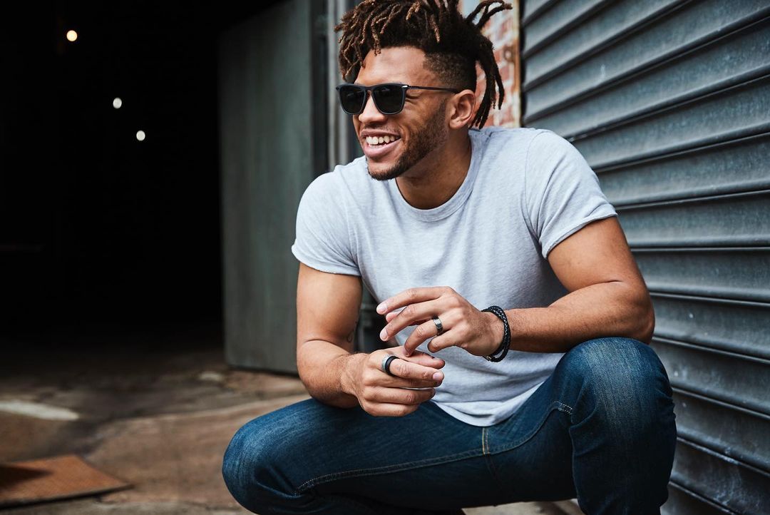 Smiling man with dreadlocks wearing sunglasses and jeans, crouching outdoors near a dark entrance, casual fashion, daytime.