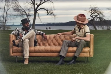 Two men seated on a tan leather tufted sofa outdoors on a lawn with a river and bare trees in the background.