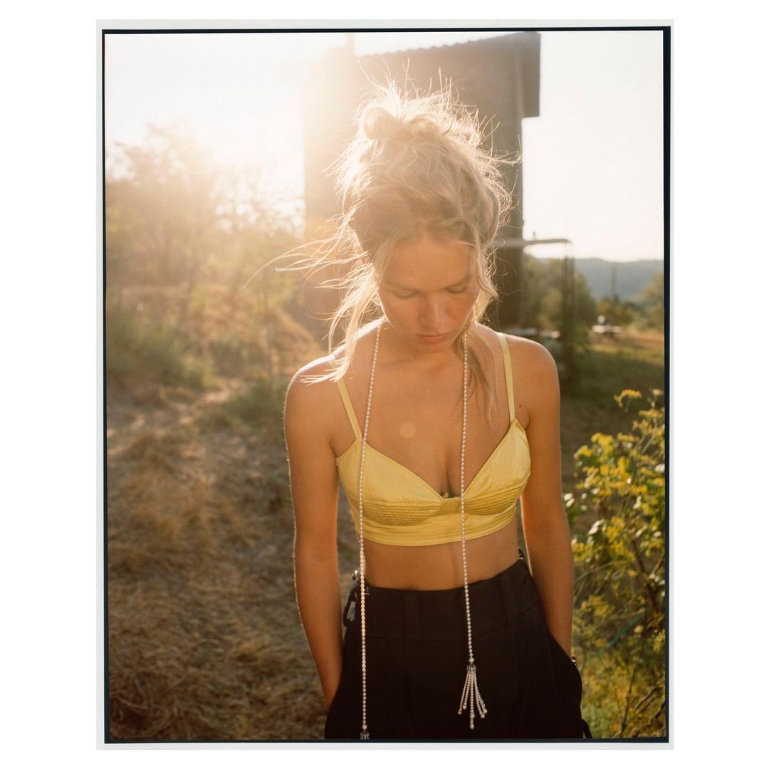 Young woman with blonde hair styled in a bun, wears a yellow bra top and dark pants, and stands outdoors in the sunlight.