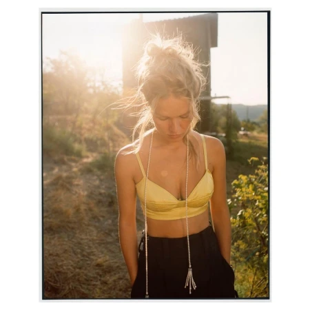 Young woman with blonde hair styled in a bun, wears a yellow bra top and dark pants, and stands outdoors in the sunlight.