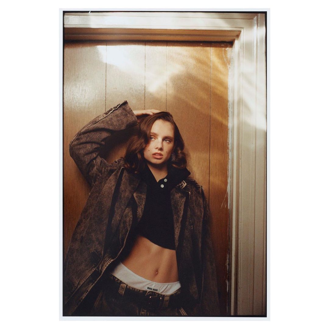 Fashion portrait of a woman in an open denim jacket, cropped hoodie, and underwear standing in a doorway with brown wood paneling.