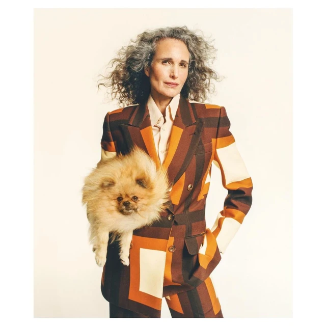 Mature woman with gray curly hair in a patterned suit holds a fluffy orange Pomeranian dog in a studio setting with a neutral background.