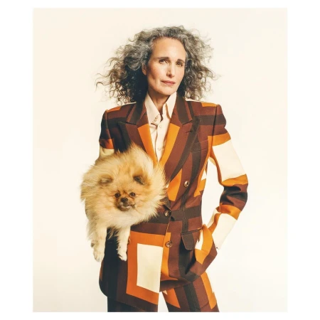 Mature woman with gray curly hair in a patterned suit holds a fluffy orange Pomeranian dog in a studio setting with a neutral background.