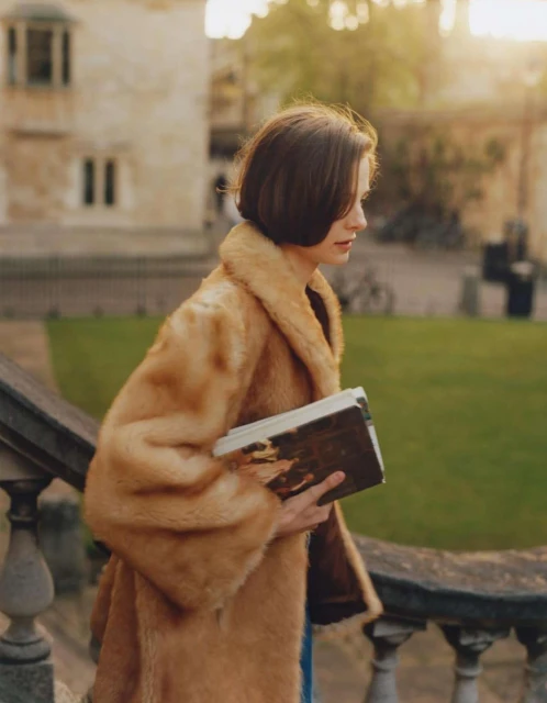 Young woman in a fur coat walks down stone steps holding books in an outdoor courtyard with grass and buildings in the background.