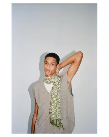Young man in a grey sweater vest wearing a patterned green scarf against a plain light wall.