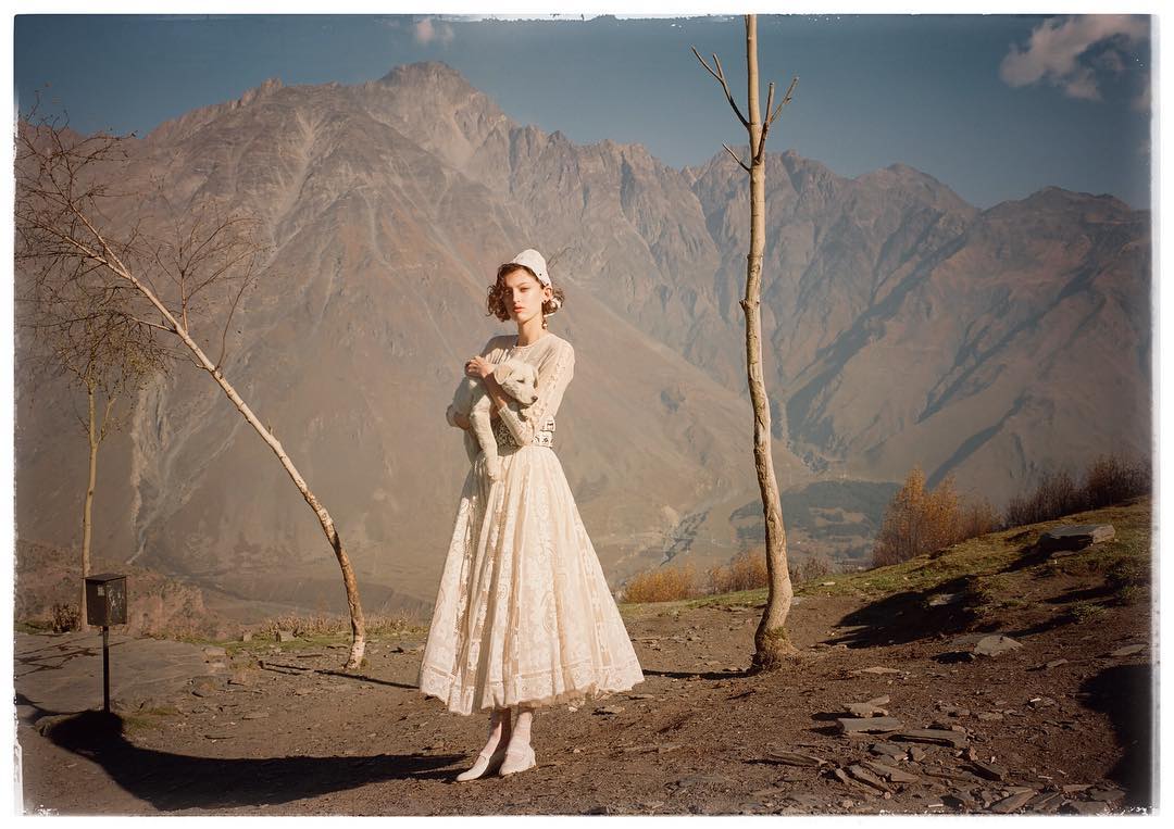 A woman in a white dress holds a white dog in a mountainous landscape. The photo captures a blend of fashion and nature, showcasing a serene, elegant scene.