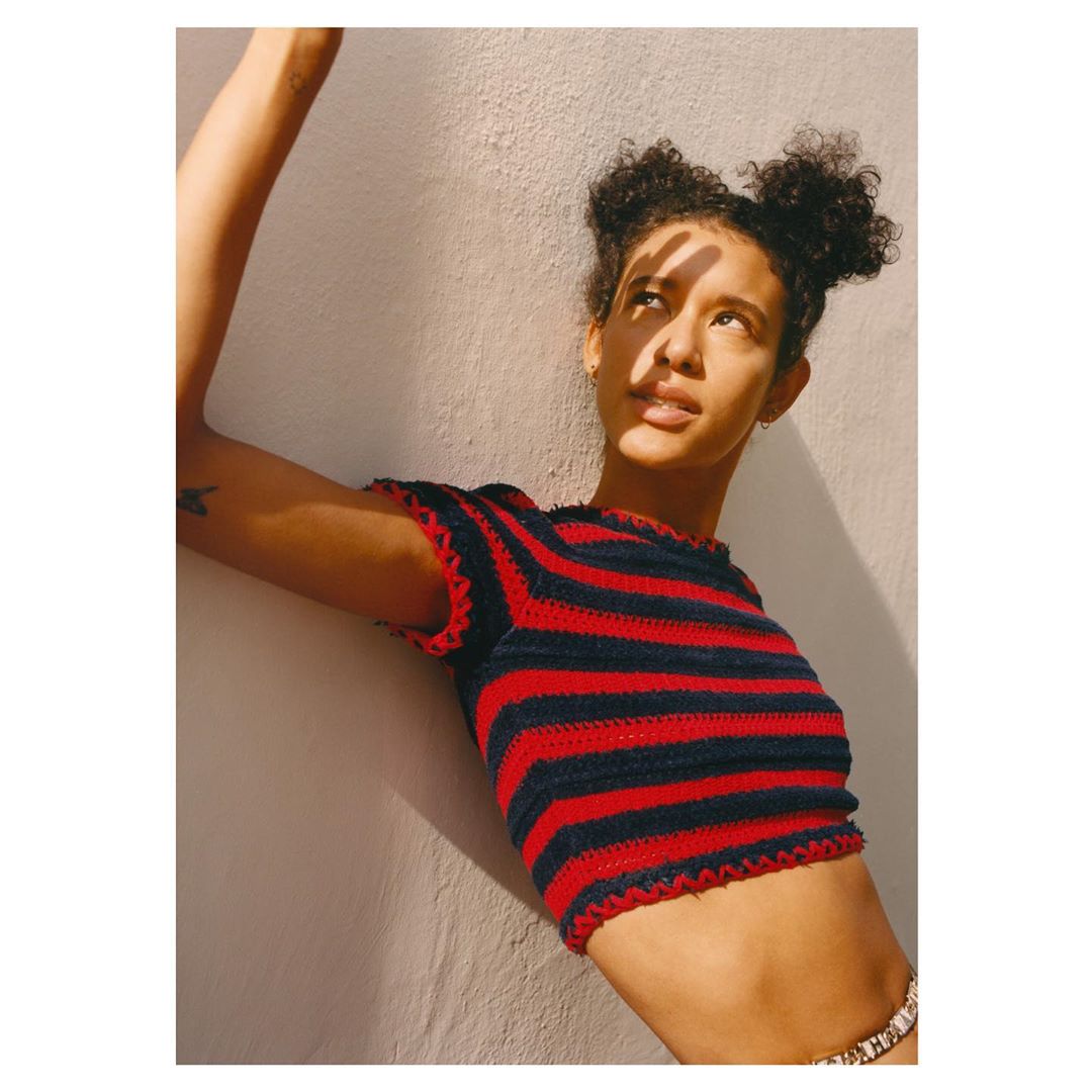Portrait of a young adult female with dark curly hair wearing a red and blue striped crop top, posing in an indoor sunlit environment.