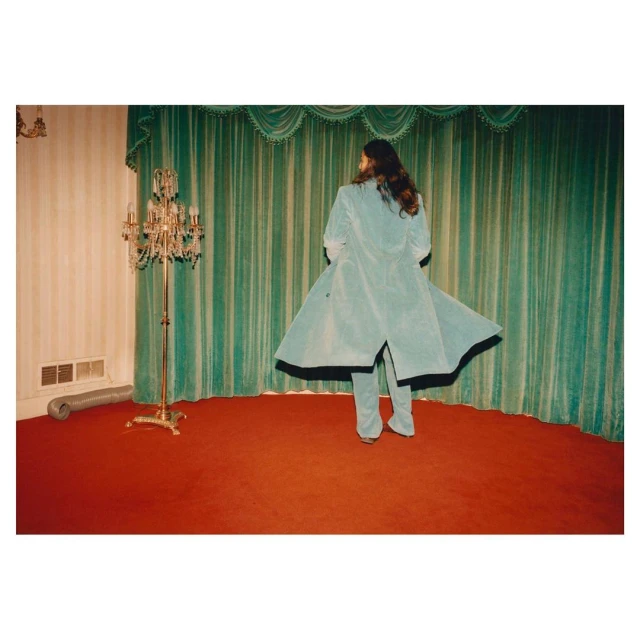 Adult female in a mint-green overcoat stands in front of a striped green curtain, on a red carpet, next to a chandelier. Fashion.