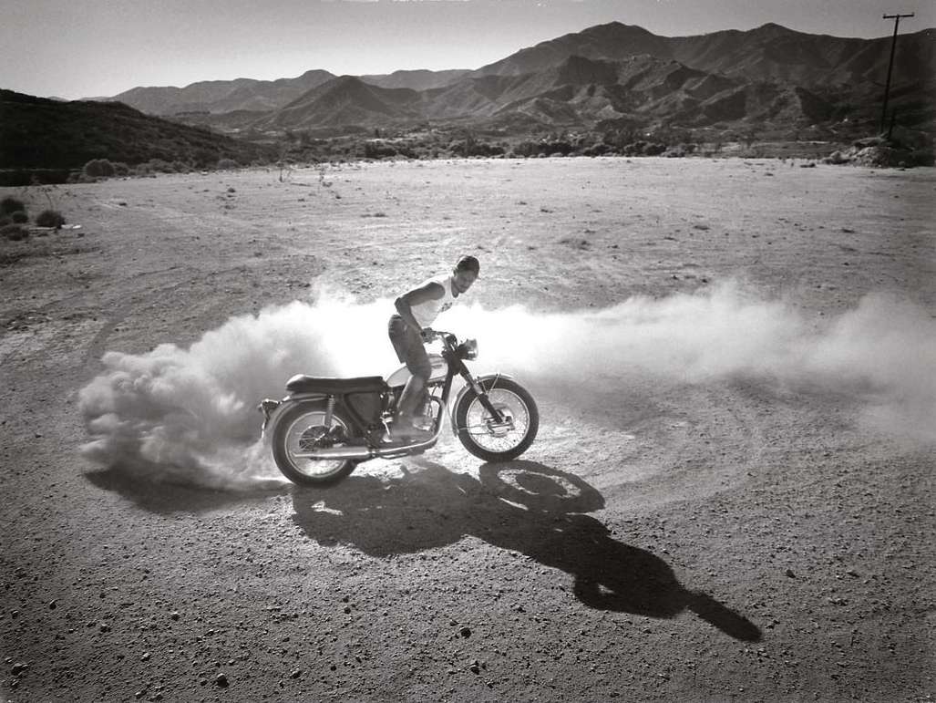 Monochrome shot of a man riding a motorcycle across a dirt field with mountains in the background, creating a cloud of dust.