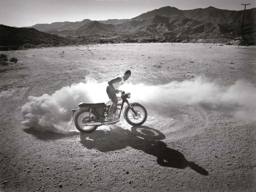 Monochrome shot of a man riding a motorcycle across a dirt field with mountains in the background, creating a cloud of dust.