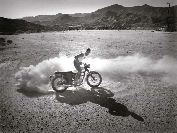 Monochrome shot of a man riding a motorcycle across a dirt field with mountains in the background, creating a cloud of dust.