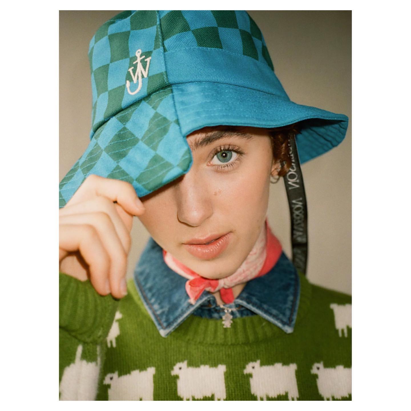 Person in a green and blue checkered bucket hat with sheep patterned sweater, posing indoors for a fashion portrait.