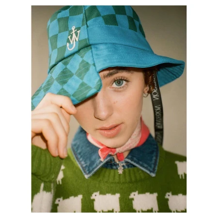 Person in a green and blue checkered bucket hat with sheep patterned sweater, posing indoors for a fashion portrait.