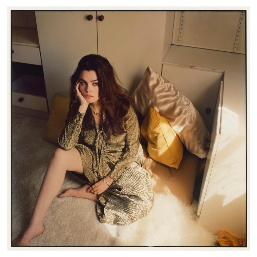Brunette woman in gold dress poses sitting on floor with pillows in casual, indoor setting with a warm, neutral color palette.