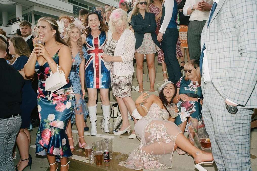 Large crowd of women in dresses and suits at an outdoor event, some sitting on steps, dressed up and laughing, socializing with drinks.