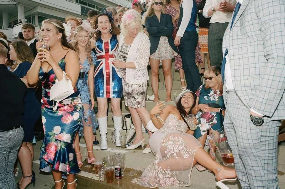 Large crowd of women in dresses and suits at an outdoor event, some sitting on steps, dressed up and laughing, socializing with drinks.