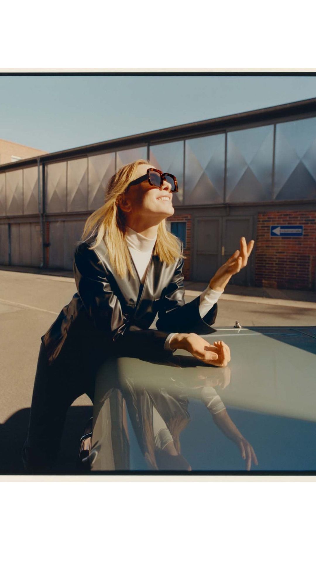 Blonde woman wearing oversized sunglasses and dark jacket leaning on a car hood outdoors under a clear sky.