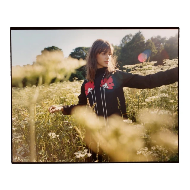 Young woman stands in a sunlit meadow with long grass and wildflowers wearing a black floral patterned sweater and looking left.
