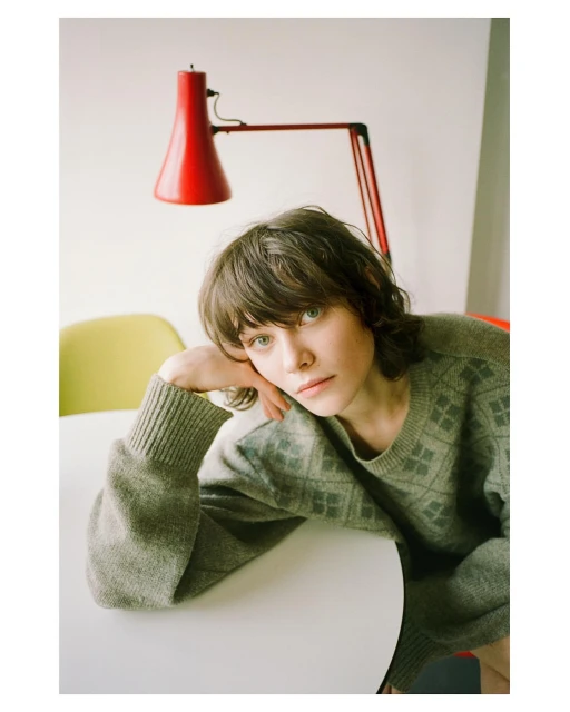 Adult female with brown hair and a grey sweater sits at a white table with a red lamp and yellow chair in the background in an indoor setting.