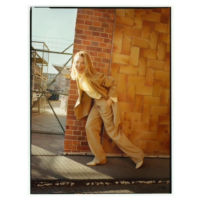Adult female in a khaki suit smiles next to a brick wall with a chain-link fence and building in the background under a blue sky.