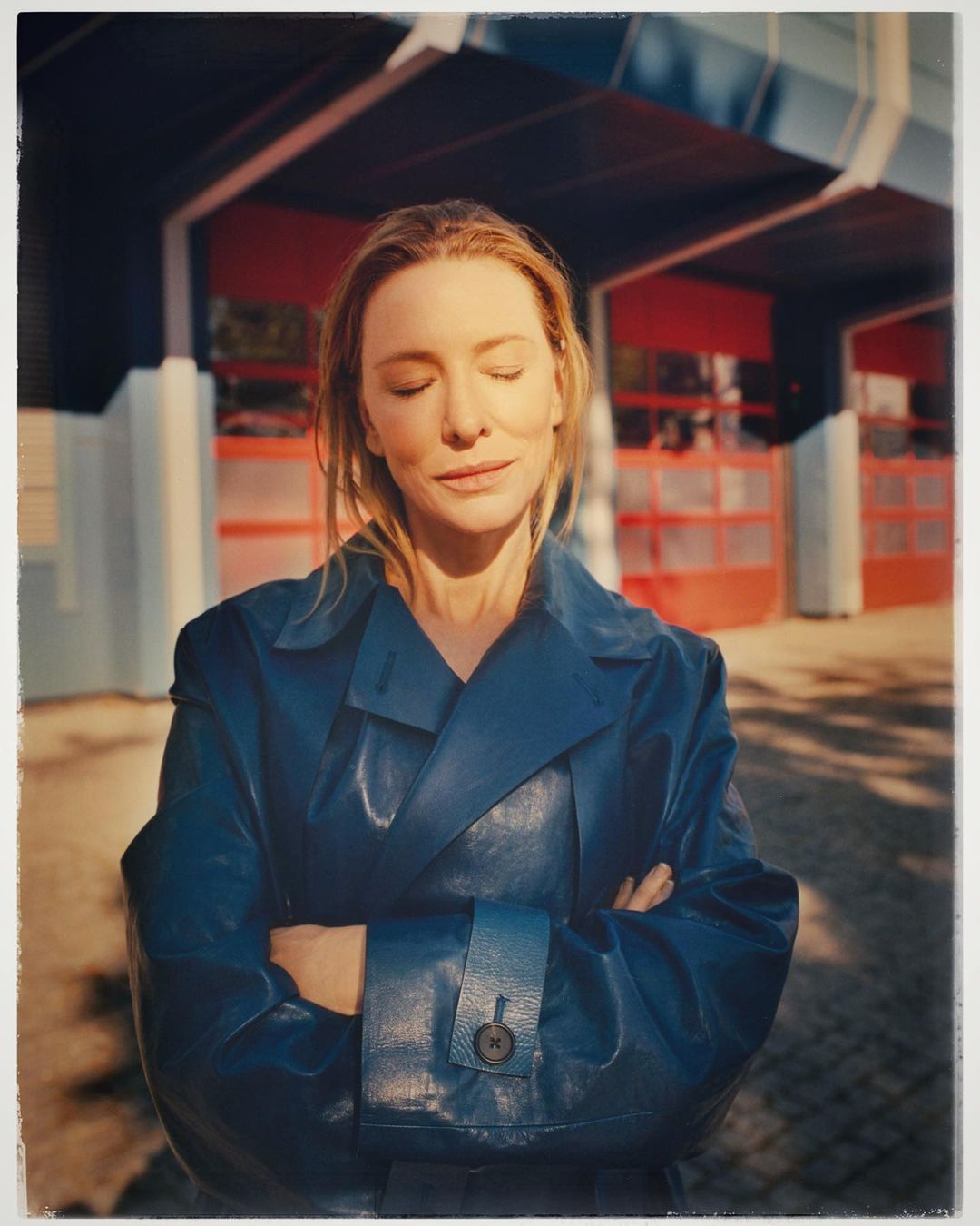 Woman in blue coat stands outdoors with eyes closed, bathed in sunlight, against a backdrop of a red and grey building.