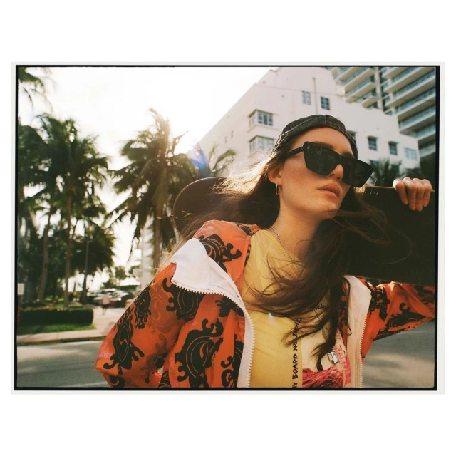 A young woman in sunglasses and a patterned jacket stands on a street lined with palm trees, holding a skateboard with a white building behind.