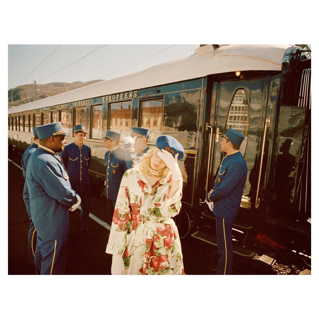 Fashion shot of a blonde woman in a floral coat with train personnel standing next to a train car with gold trim and dark windows.