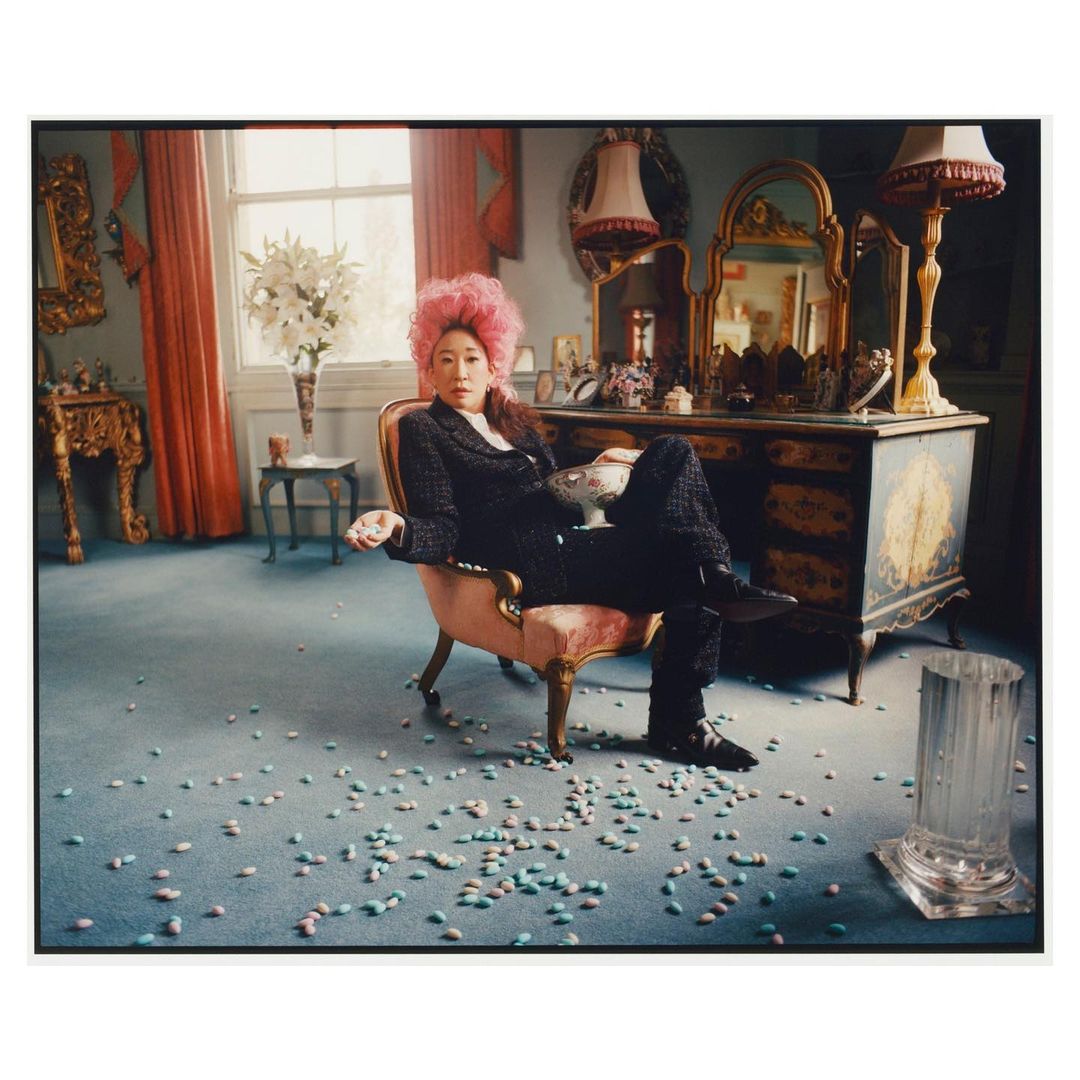 Elegant woman with pink hair poses in ornate room with scattered candies, showcasing fashion and editorial style.