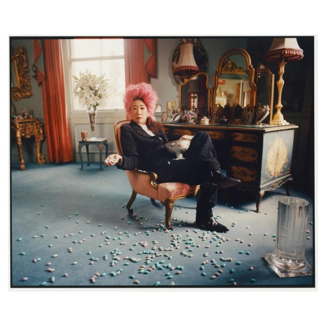 Elegant woman with pink hair poses in ornate room with scattered candies, showcasing fashion and editorial style.