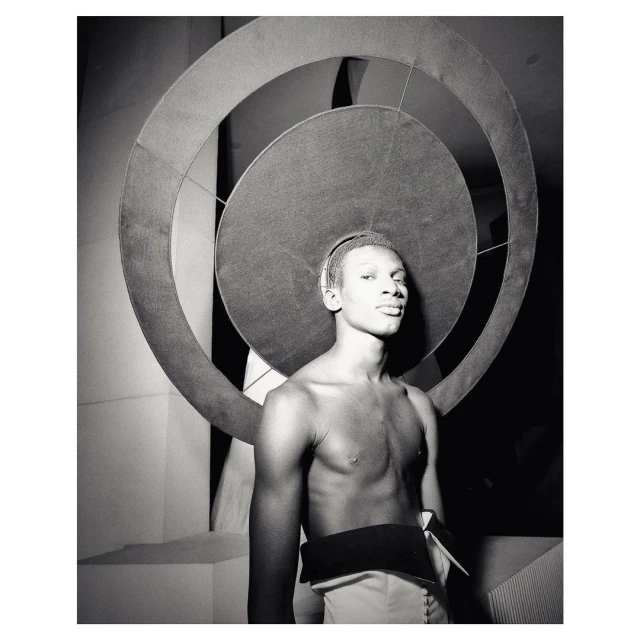 Striking portrait of a topless person wearing a large circular headpiece, standing in front of geometric shapes, black and white.