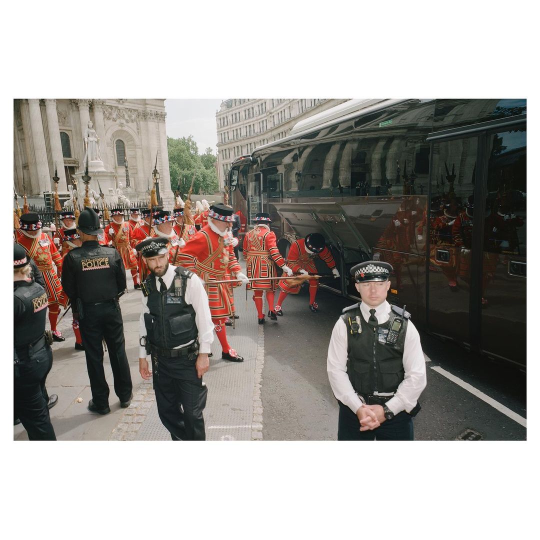 Guards in traditional red uniforms and police officers in black uniforms stand on a city street near a tour bus and building.