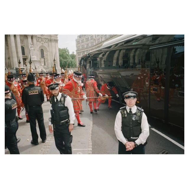 Guards in traditional red uniforms and police officers in black uniforms stand on a city street near a tour bus and building.